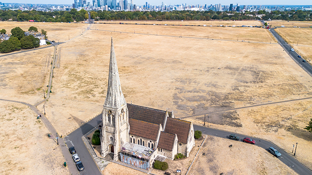 Aerial view of a church surrounded by parched yellow-brown fields with a city skyline in the distance, highlighting widespread drought and dry grass under clear summer skies.