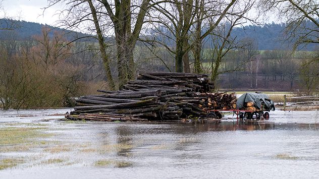 Holzstapel und Anhänger stehen in einer überfluteten Wiese. Kahle Bäume und grauer Himmel im Hintergrund. (c) dpa