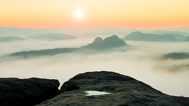 Nebelige Hügellandschaft bei Sonnenaufgang, Blick von einer Felsplatte mit Pfütze im Vordergrund.