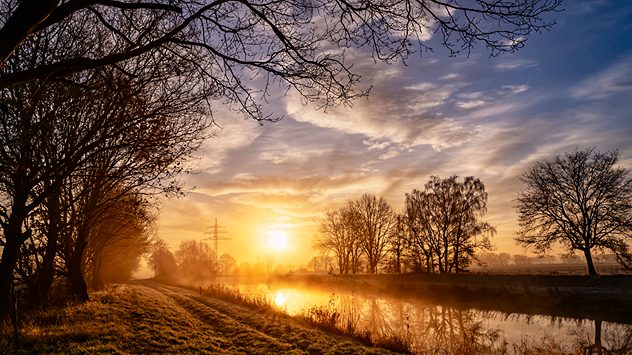 Sonnenaufgang über einem Fluss im Winter mit Nebel und kahlen Bäumen am Ufer. (c) Petra Quade