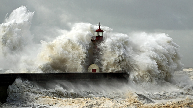 Ein gewaltiger Sturm trifft auf die Küste – haushohe Brecher schlagen mit voller Wucht gegen einen Leuchtturm auf der Mole. Die tosende See, peitschender Wind und aufgewühlte Gischt machen die Urgewalt des Unwetters eindrucksvoll sichtbar.