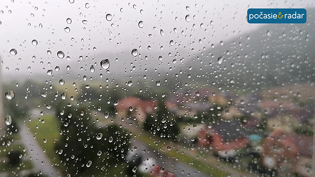 Dažďové kvapky na okne s rozmazaným výhľadom na obec a zamračenú krajinu v pozadí