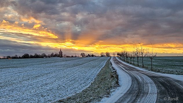 Verschneite Landstraße zwischen Feldern bei Sonnenuntergang. Dunkle Wolken und orangefarbenes Abendlicht am Horizont. (c) Daniela BÃ¶hme