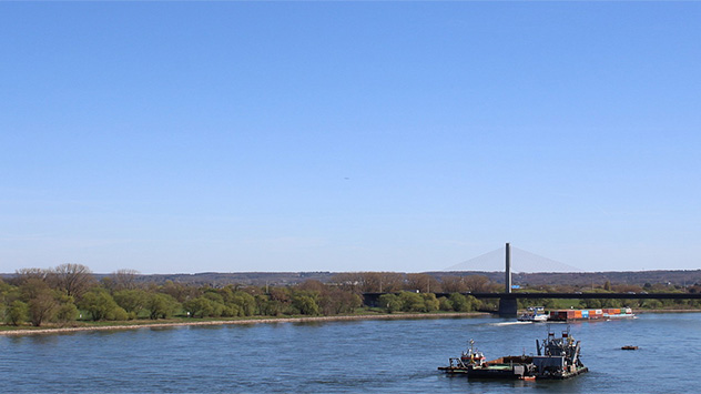 Flusslandschaft am Rhein in Bonn bei Sonnenschein mit Arbeitsschiff und Brücke im Hintergrund.