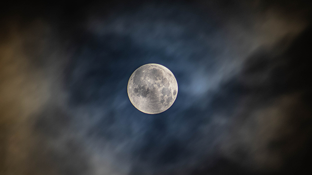 Bright full moon centred in a dark night sky surrounded by wispy clouds with blue and golden hues.