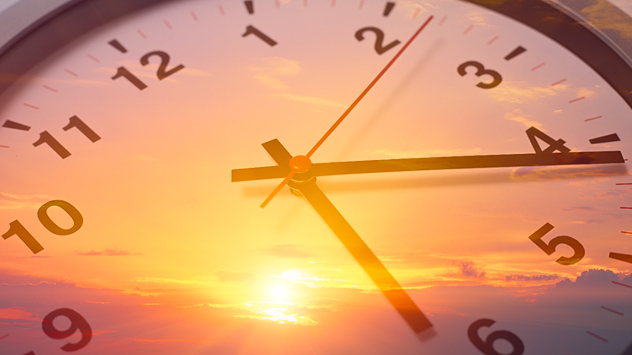 Close-up of a clock face overlaid on a golden sunrise sky, symbolizing time change and longer daylight hours.