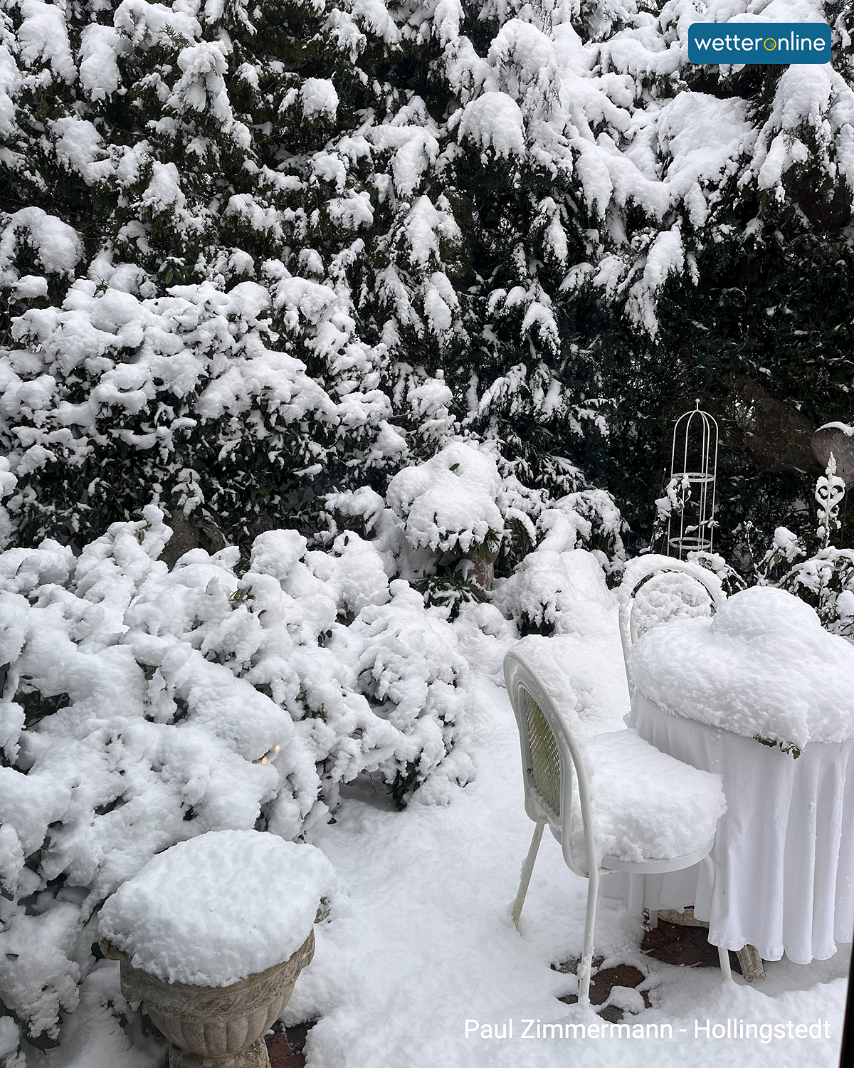 Verschneiter Gartentisch und Stühle zwischen schneebedeckten Sträuchern.