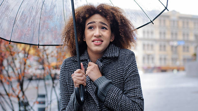 Junge Frau mit Regenschirm und Wintermantel friert im herbstlichen Regen.