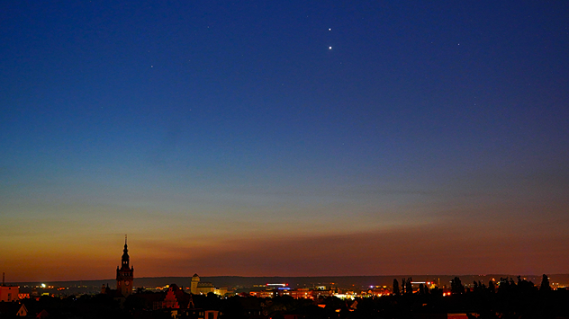 Abendhimmel über einer Stadt mit Kirchen und Lichtern. Zwei helle Planeten leuchten am dunkler werdenden Himmel.