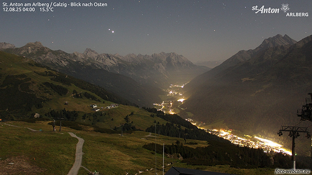 Blick auf eine erleuchtete Bergregion bei Nacht. Über den Bergen ein klarer Sternenhimmel mit sichtbaren Sternbildern.