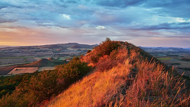 Von warmem Abendlicht rot und orange beleuchteter Hügelkamm mit trockenem Gras und Sträuchern; im Hintergrund erstrecken sich Felder, sanfte Berge und ein blau-grauer Wolkenhimmel.