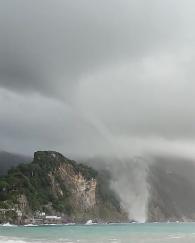 Tornado over the sea near a coastline beneath dark clouds.