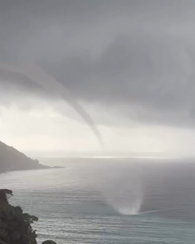 Tornado over the sea near a coastline beneath dark clouds.