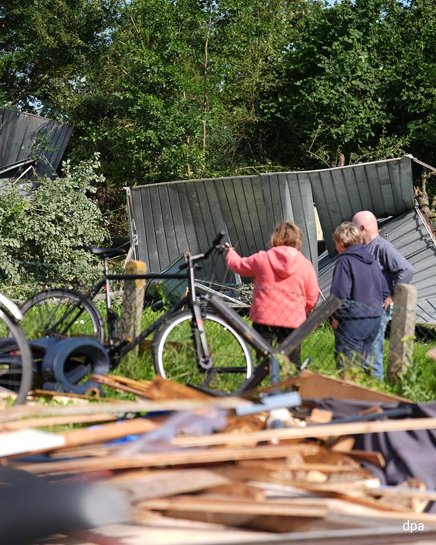 Drei Menschen stehen vor den Trümmern zerstörter Garagen und Gartenlauben. Im Vordergrund liegen Holz, Dachziegel und weitere Trümmerteile auf einem Grundstück.