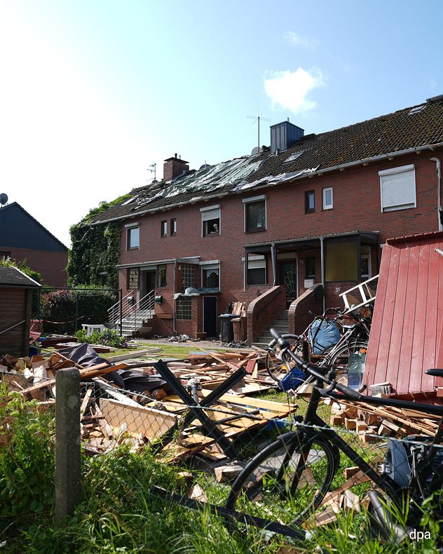 Reihenhäuser mit provisorisch abgedeckten Dächern nach einem Tornado. Im Vordergrund liegen Trümmer einer Gartenlaube, Holz und Dachziegel verstreut auf einem Grundstück.