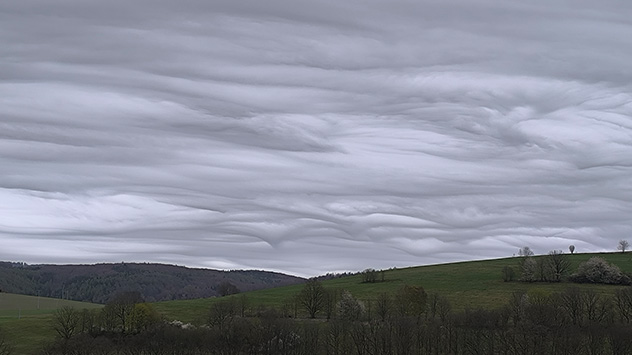 Wellenartige Wolkenformation über einer hügeligen Landschaft mit Bäumen.