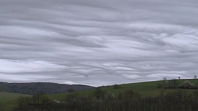 Wellenartige Wolkenformation über einer hügeligen Landschaft mit Bäumen.