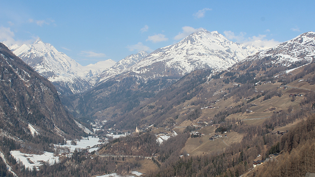 Das Webcambild zeigt ein Tal in den Alpen. Im Tal liegt noch Schnee, die Hänge sind hingegen grün.