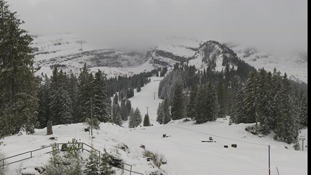 Verschneite Berglandschaft mit Skipiste und dunklen Tannen unter grauem Himmel. Die Gipfel im Hintergrund liegen teils in Wolken und Nebel.