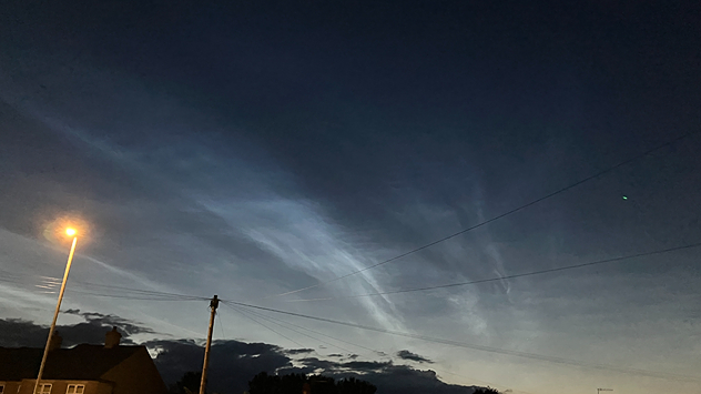Wispy electric-blue noctilucent clouds glowing above a dark twilight horizon, with rooftops, utility poles, streetlights, and overhead wires silhouetted beneath the night sky.
