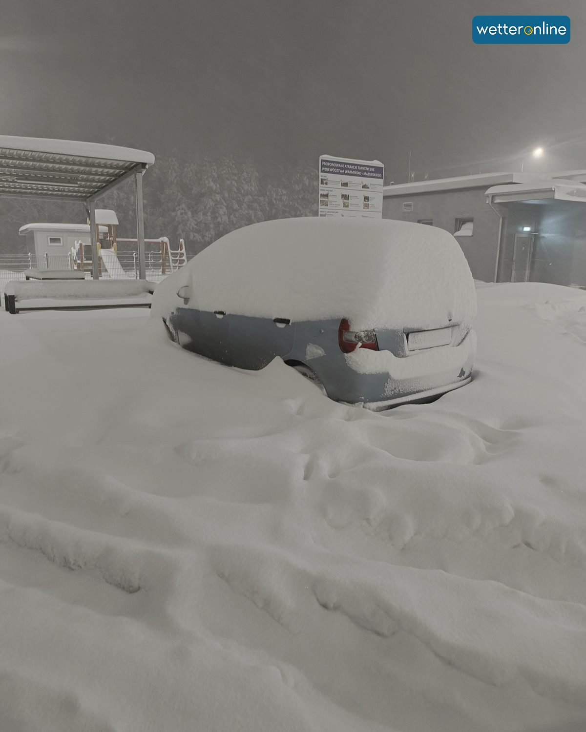 Car completely covered in snow in a car park at night. Thick fresh snow covers the vehicle and ground.