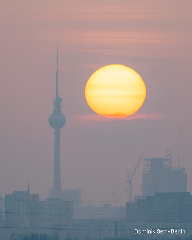 Große tiefstehende Sonne über der Skyline von Berlin mit Fernsehturm. Dunstige Luft lässt Gebäude und Kräne im Hintergrund verschwimmen.