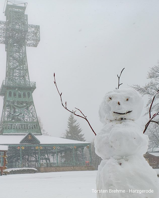 Schneemann steht im dichten Schneefall vor einem Förderturm im Harz. Alles ist weiß bedeckt, Sicht durch Nebel eingeschränkt.