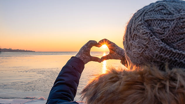 Eine Frau formt mit ihren Händen ein Herz, durch das die Wintersonne scheint.
