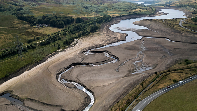 Aerial view of a depleted reservoir with exposed banks and a narrow winding stream through the dry basin, surrounded by green hills and roads.