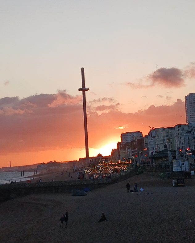 Orange sunset glowing behind a seaside city skyline, with dark clouds, a tall observation tower, beachfront buildings and people along the shore.
