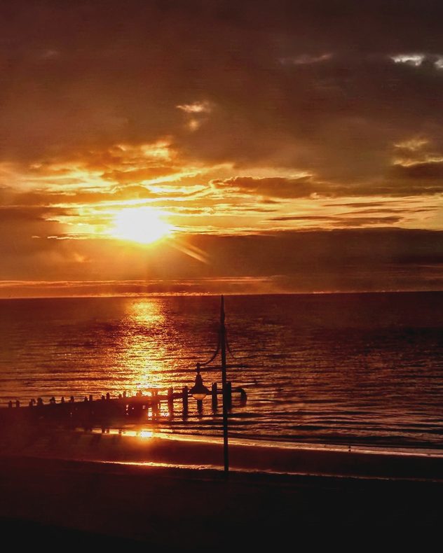 Golden Sun setting through layered clouds over the sea, casting a bright reflection across the water toward a silhouetted pier and beachfront lamps.