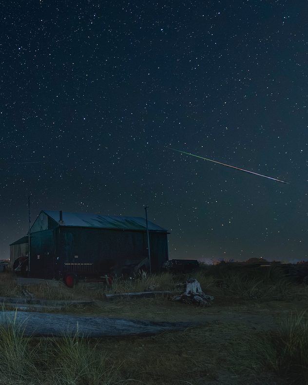 Bright multicoloured meteor streaks across a densely star-filled night sky above a dark rural building, grasses and fallen logs.