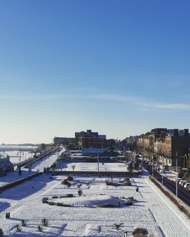 Elevated view of a coastal town blanketed in snow, with a formal park in the foreground, snow-covered streets and rooftops, terraced buildings to the right, and a clear blue sky overhead.