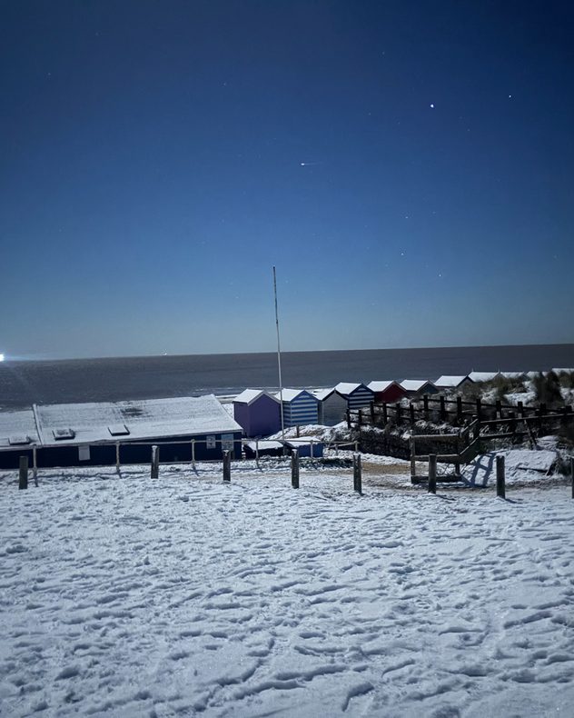 Snow-covered beach and promenade at night with a row of colorful beach huts, footprints in the snow, calm sea on the horizon, and a clear dark blue sky dotted with stars.