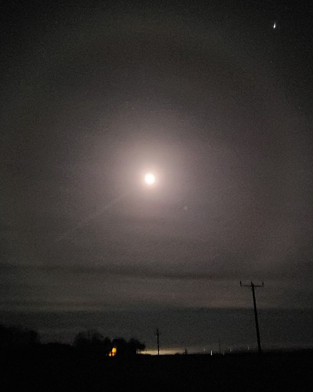 Bright full moon surrounded by a soft circular halo in a dark, hazy night sky, with faint stars visible and silhouettes of distant trees, rooftops, and utility poles along the horizon.