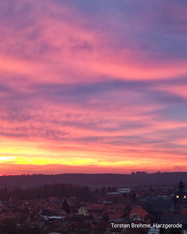 Farbenprächtiger Abendhimmel in Rot und Violett über einer Kleinstadt. Silhouetten von Dächern und Bäumen heben sich vor dem leuchtenden Horizont ab.