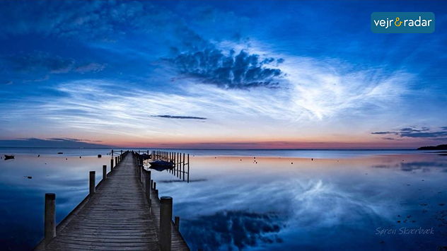 Natlysende skyer på himlen over stille vand med træbro ved solnedgang.