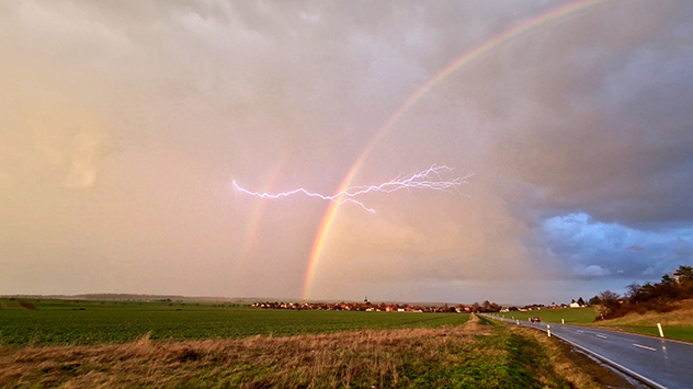 Gewitterwolke spukt Blitz über Feld aus, gleich zwei Regenbögen zu sehen.