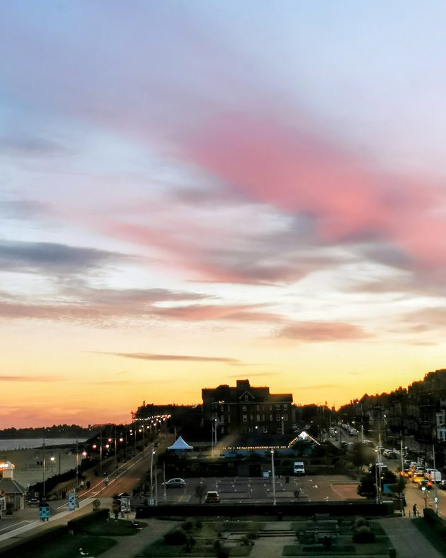 Soft pink and lavender clouds stretch across a golden sunset sky above a coastal town, with seafront buildings, roads, and glowing streetlights.