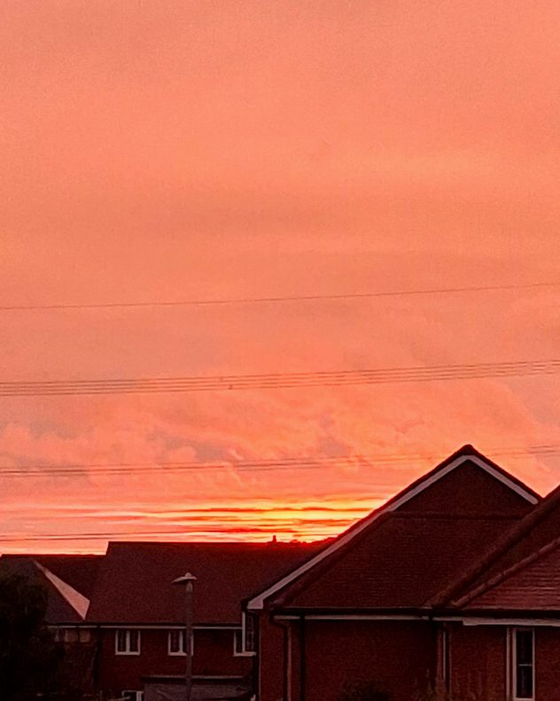 Vivid orange-red sunset fills the sky above the dark suburban rooftops, with glowing horizontal cloud bands and power lines crossing.