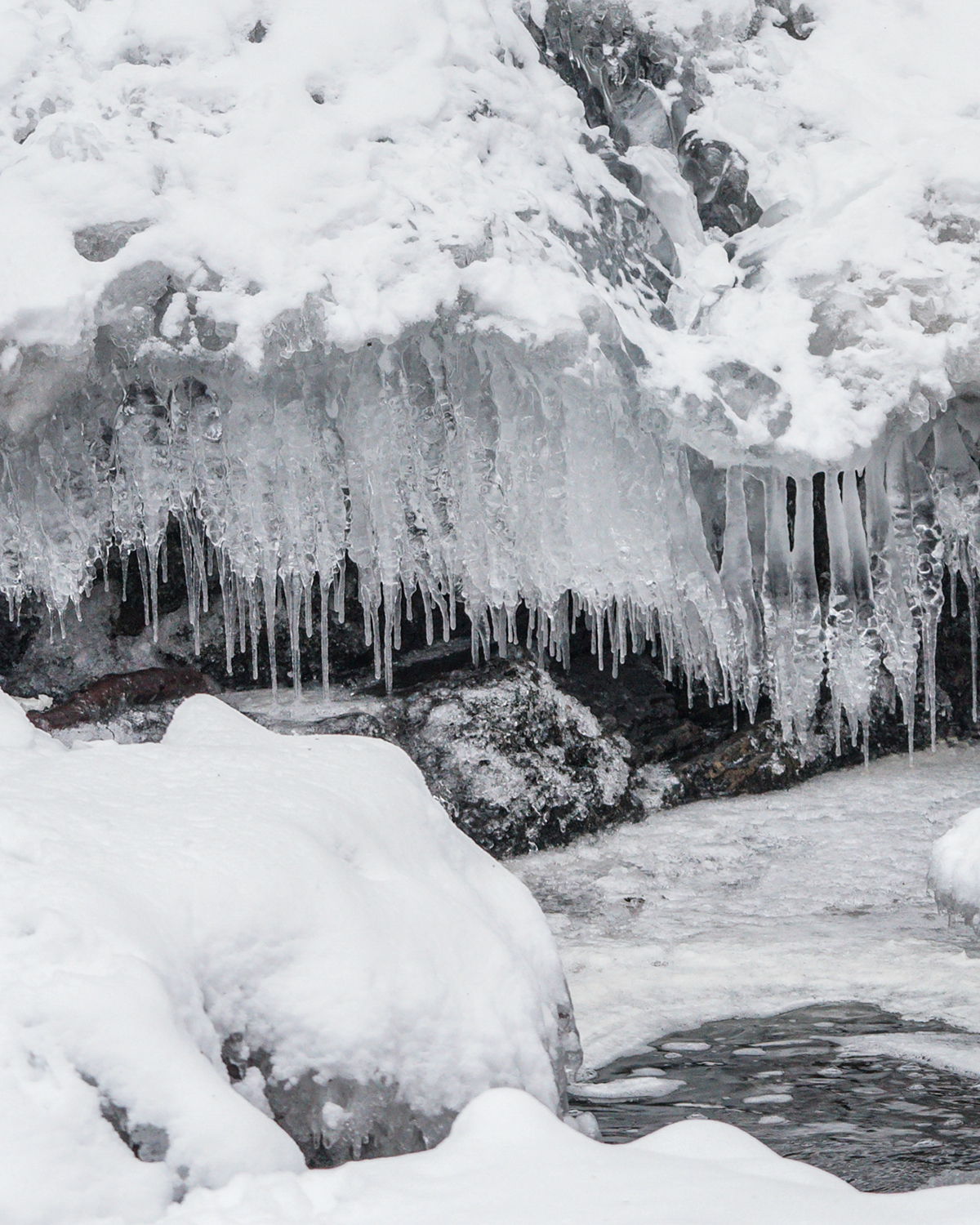 Close-up of long clear icicles hanging from a snow-covered rock ledge above partially frozen water, with layered ice, snow, and dark stone visible.