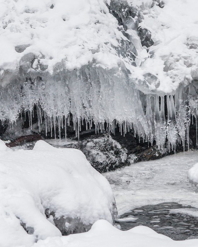 Close-up of long clear icicles hanging from a snow-covered rock ledge above partially frozen water, with layered ice, snow, and dark stone visible.