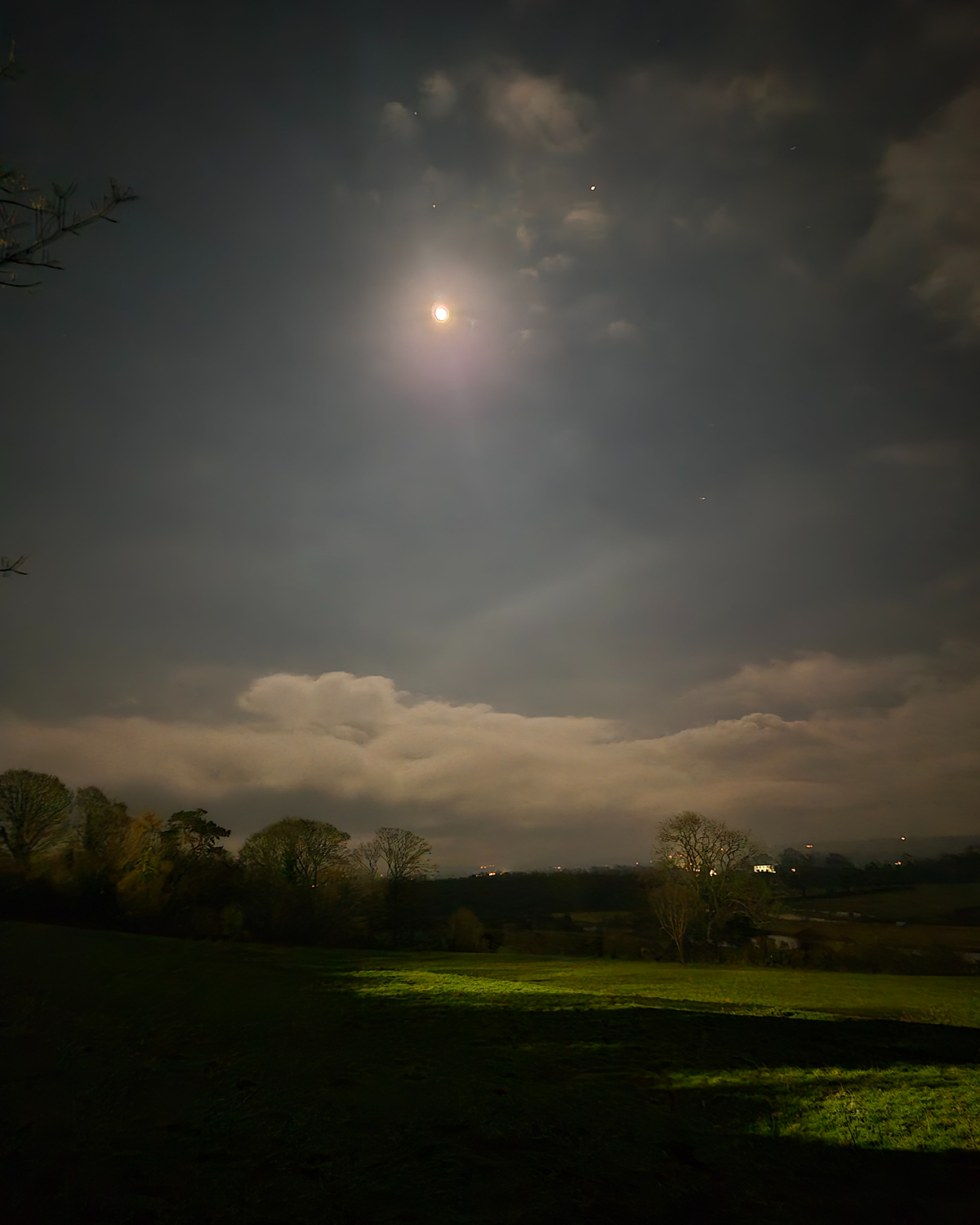 Bright moon with a soft circular halo in a cloudy night sky above rolling fields and tree silhouettes, with faint stars and distant lights on the horizon.