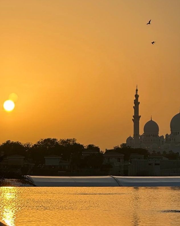 Golden sunset over calm water with the sun low on the horizon, silhouetted mosque domes and a tall minaret on the right, and two birds flying across an orange sky.