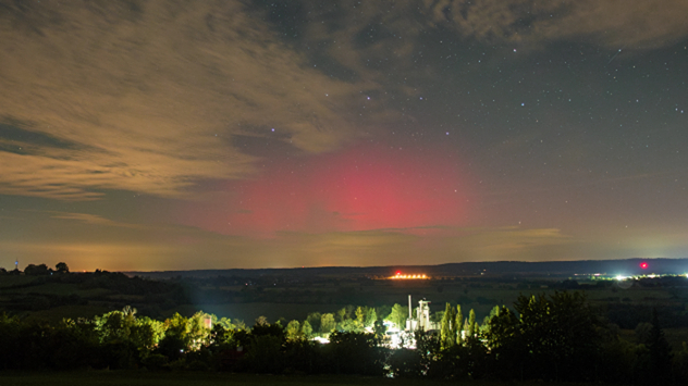Rotes Polarlicht über süddeutscher Landschaft, mit erleuchteten Gebäuden und teils wolkigem Nachthimmel.