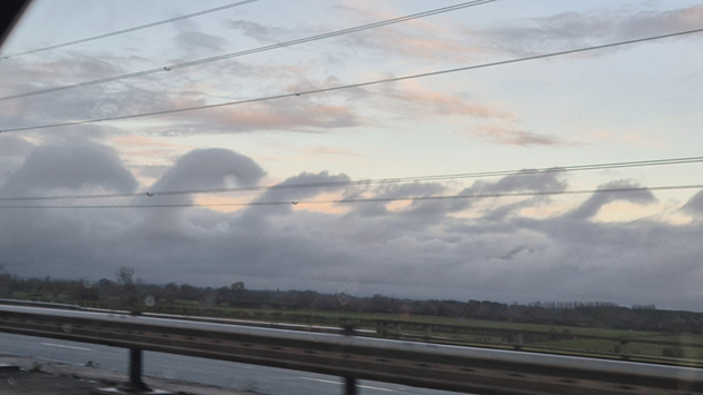 Wave-shaped Kelvin-Helmholtz clouds forming above the England–Scotland border, viewed from a roadside with railings and power lines.