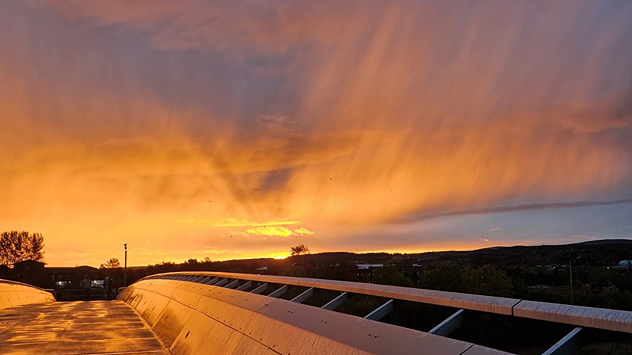 Golden-orange sunset over Inverness with streaked clouds and rain bands, reflected on the curved surface of a modern bridge.