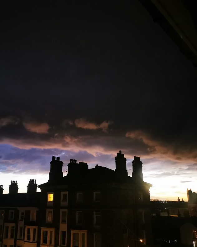 Dark storm clouds loom over silhouetted rooftops and chimneys at sunset, with a glowing orange horizon and a lit window in one building.