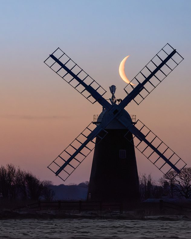 Silhouette of a traditional windmill at dusk with a crescent moon positioned between its sails against a soft purple and orange twilight sky.
