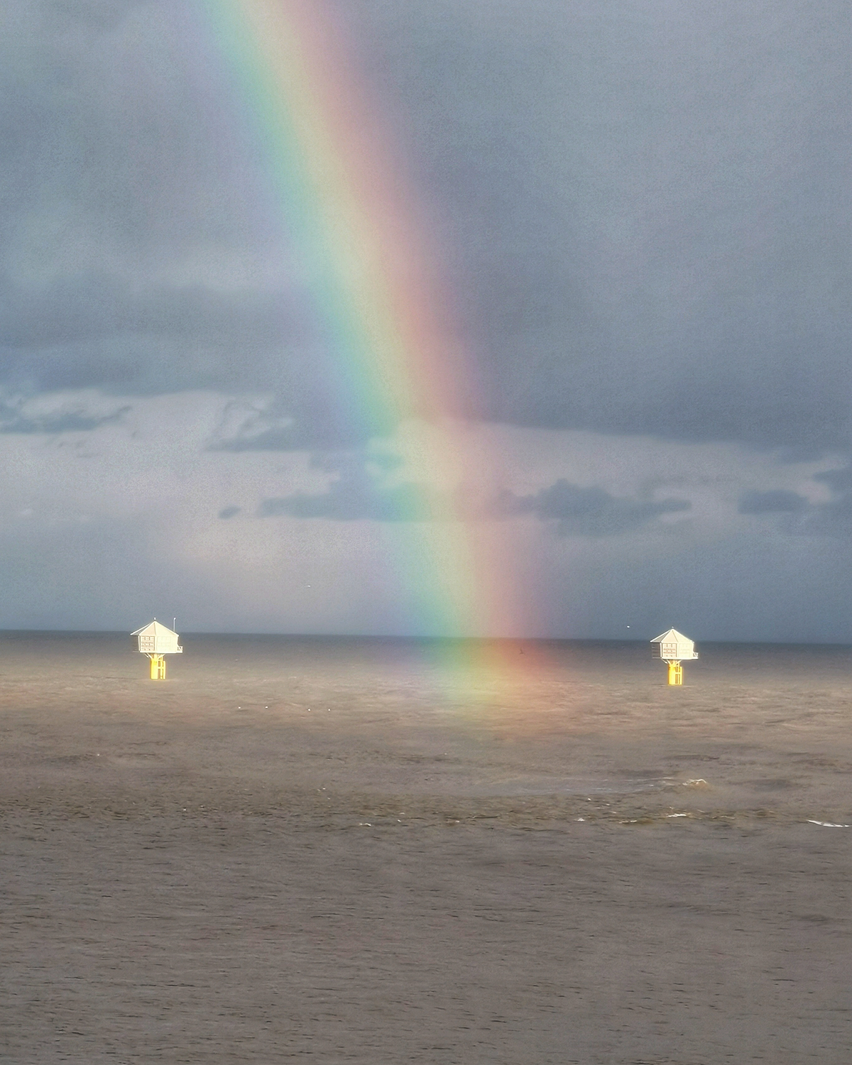 Bright rainbow descending into the sea under dark storm clouds, with two offshore structures visible on the water.